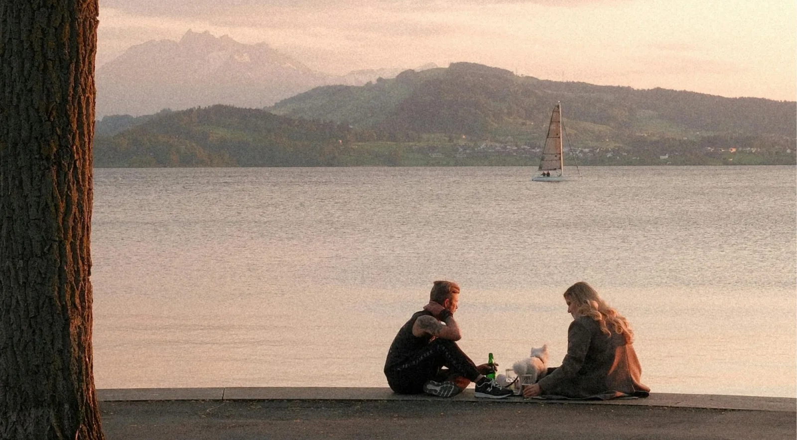 A couple enjoying a lakeside picnic at sunset with a sailboat on the water and mountains behind