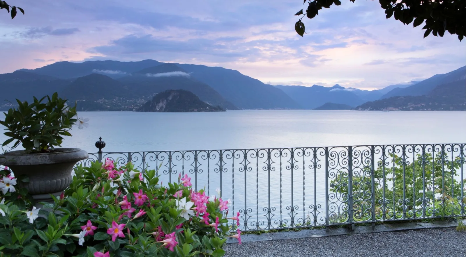 A villa terrace at dusk with a wrought-iron railing and pink flowers overlooking Lake Como and the mountains