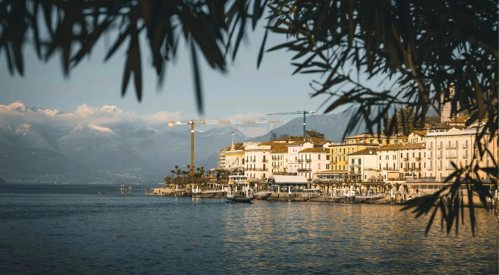 Bellagio's waterfront at golden hour, framed by leaves with snow-capped mountains behind